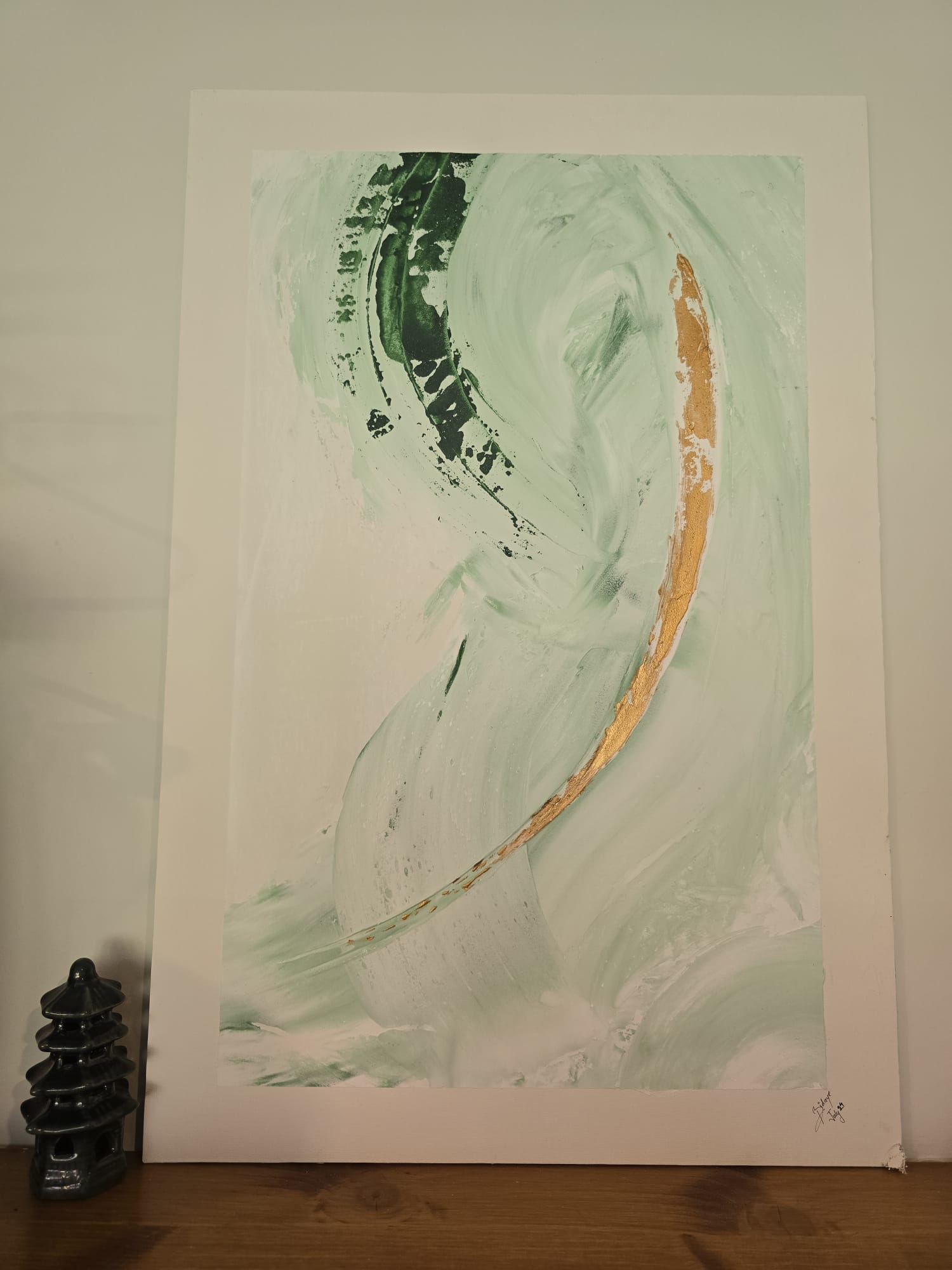 An abstract painting on white board, signed by Mr Bidaye and propped on a wooden shelf beside a small green pagoda ornament: broad pale mint brushstrokes swirling upward, a dark forest green sweep at the top left, and a single curved stroke of gold leaf running down the right side.